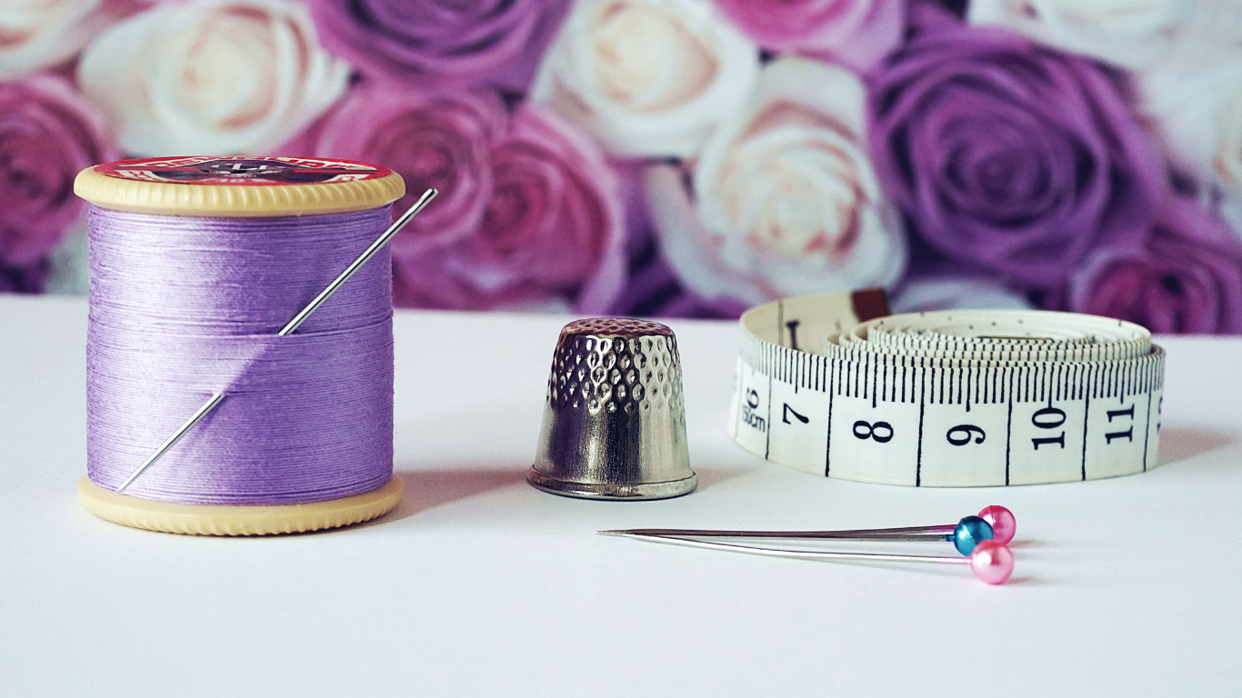 Spool of purple thread, thimble, tape measure, and pins on a white surface with a floral background.